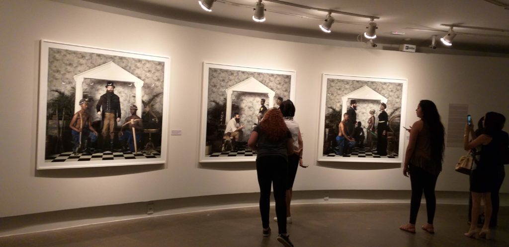 Fotografia da exposição ExAfrica com 3 quadros grandes alinhados lado a lado e algumas pessoas apreciando. 