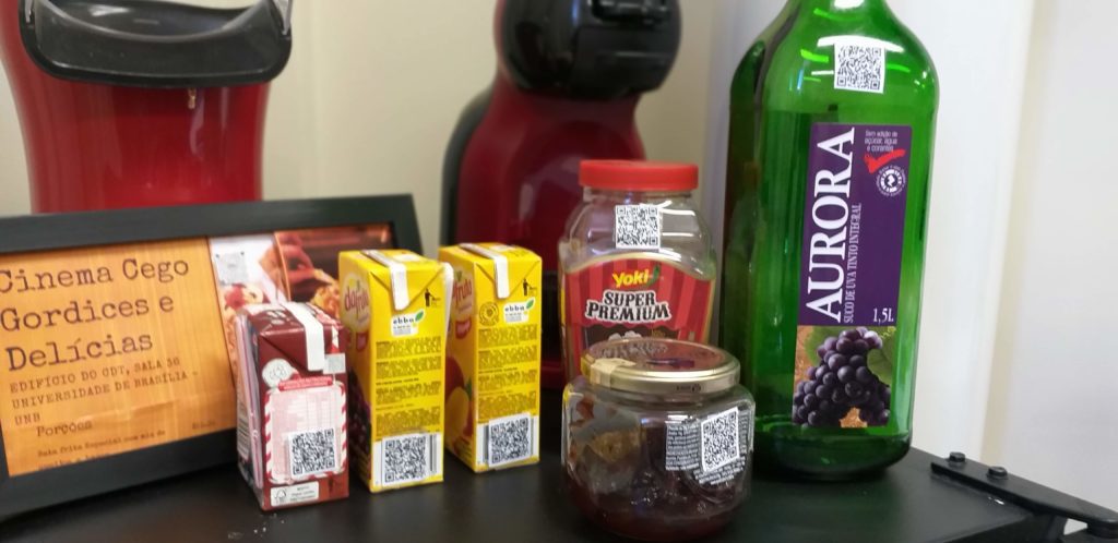 Fotografia aproximada de alguns produtos de consumo identificados com qrcode de audiodescrição. Da esquerda pra a direita, uma plaquinha escrito Cinema Cego, gordices e delícias acompanhada de uma caixinha pequena de suco de uva, suco de manga, um toddynho, um pote de geleia, pote de milho de pipoca e garrafa de vidro com suco de uva.