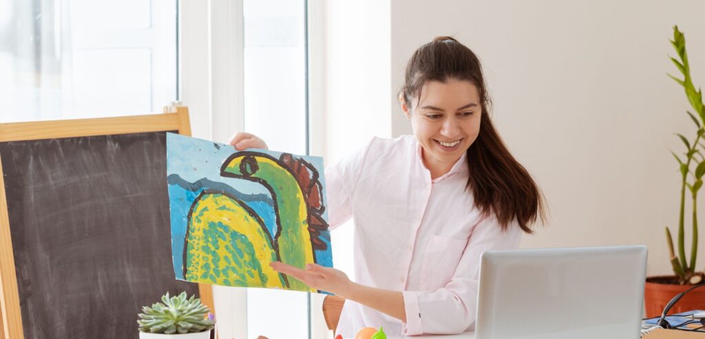 Fotografia colorida de uma professora segurando um cartaz com o desenho de um dinossauro, à frente do computador. 