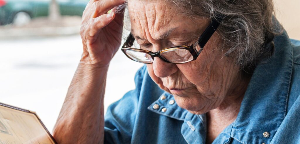 Fotografia colorida de uma idosa lendo um cardápio. Ela tem aproximadamente 70 anos, usa óculos e está com a mão direita na cabeça, em sinal de preocupação.