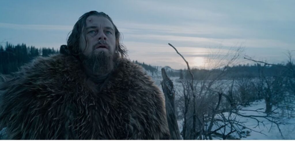 Cena de O Regresso, filme de 2015. Filtro azul para destacar o Frio, isolamento e melancolia do personagem em cena, interpretado pelo ator Leonardo Di Caprio. Ele está um ambiente cercado de neve e usa um grande casaco de pele. Ao fundo, céu azul corta a linha do horizonte.
