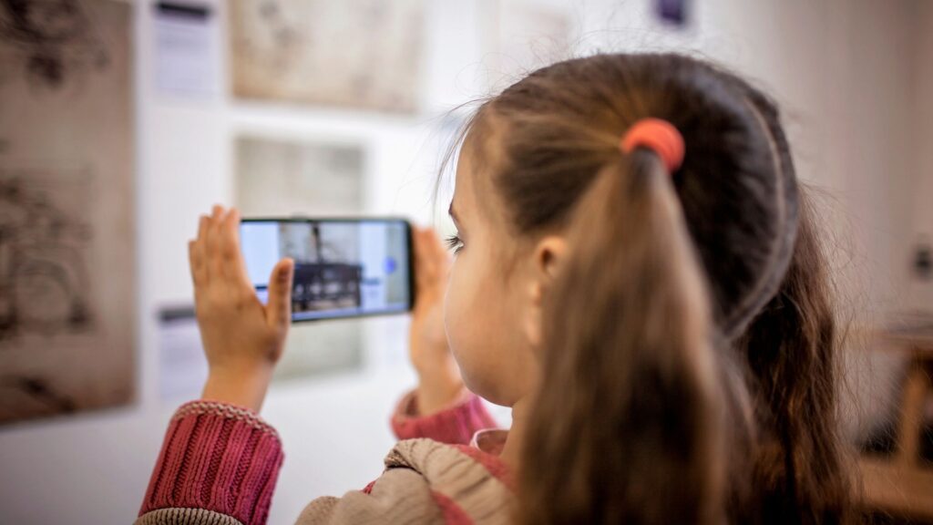fotografia de uma garota em uma exposição de arte. Ela segura o celular em frente aos olhos e o direciona para um quadro, exposto na parede. 
