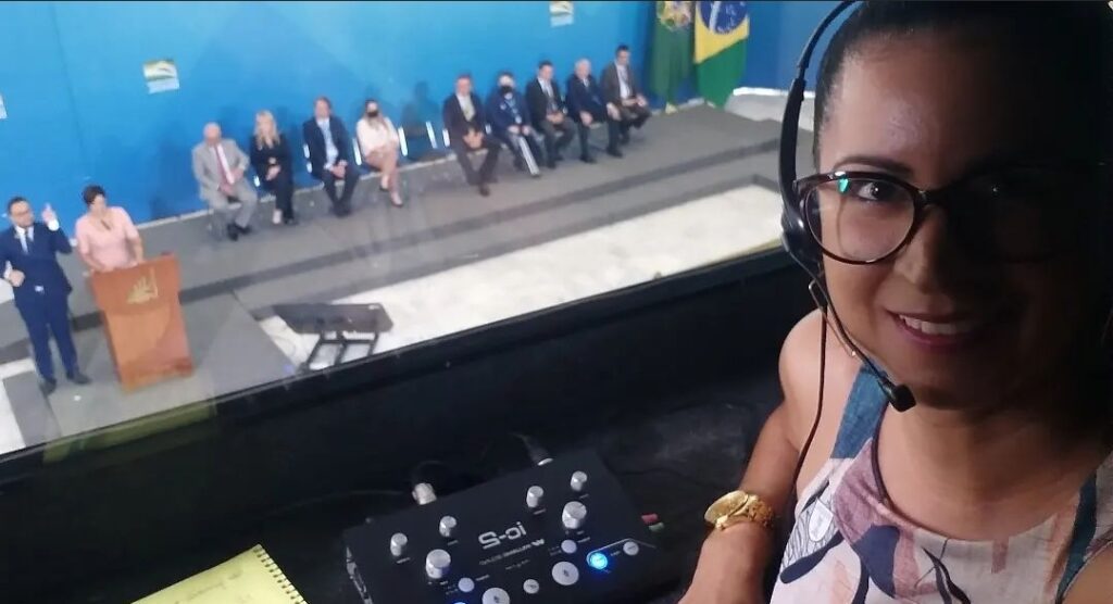 Fotografia de cerimônia realizada na presidência da república com a primeira dama, Michele Bolsonaro realizando discurso. Ao lado dela, um intérprete de Libras. Ao fundo, sentados lado a lado, o presidente jair Bolsonaro e outras autoridades. 