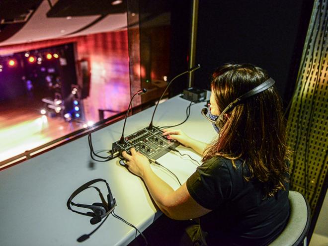 Fotografia de uma mulher dentro de uma cabine acústica. Ela está sentada e apoia os braços na mesa, onde estão localizados os microfones de transmissão e rádios receptores.